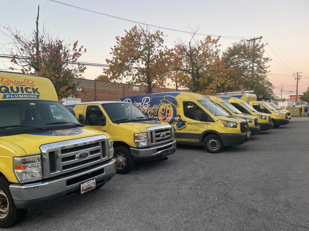 image2 1 - Brian B. Quick Yellow Brian Quick Plumbing vans parked in a lot, visible logos on the sides and front; autumn trees and power lines in the background.
