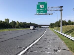 Green highway exit sign for Route 216 with 'Fulton' and 'Laurel' visible, arrows pointing toward the exit, over a multi-lane road with cars ahead and an overpass in the distance.
